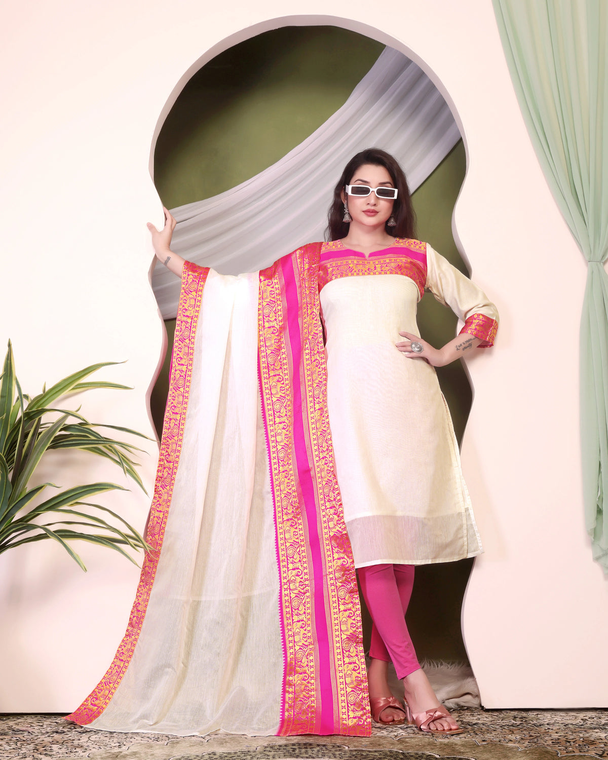 Woman wearing a white and pink traditional outfit with a decorative dupatta, standing in front of a decorative archway.