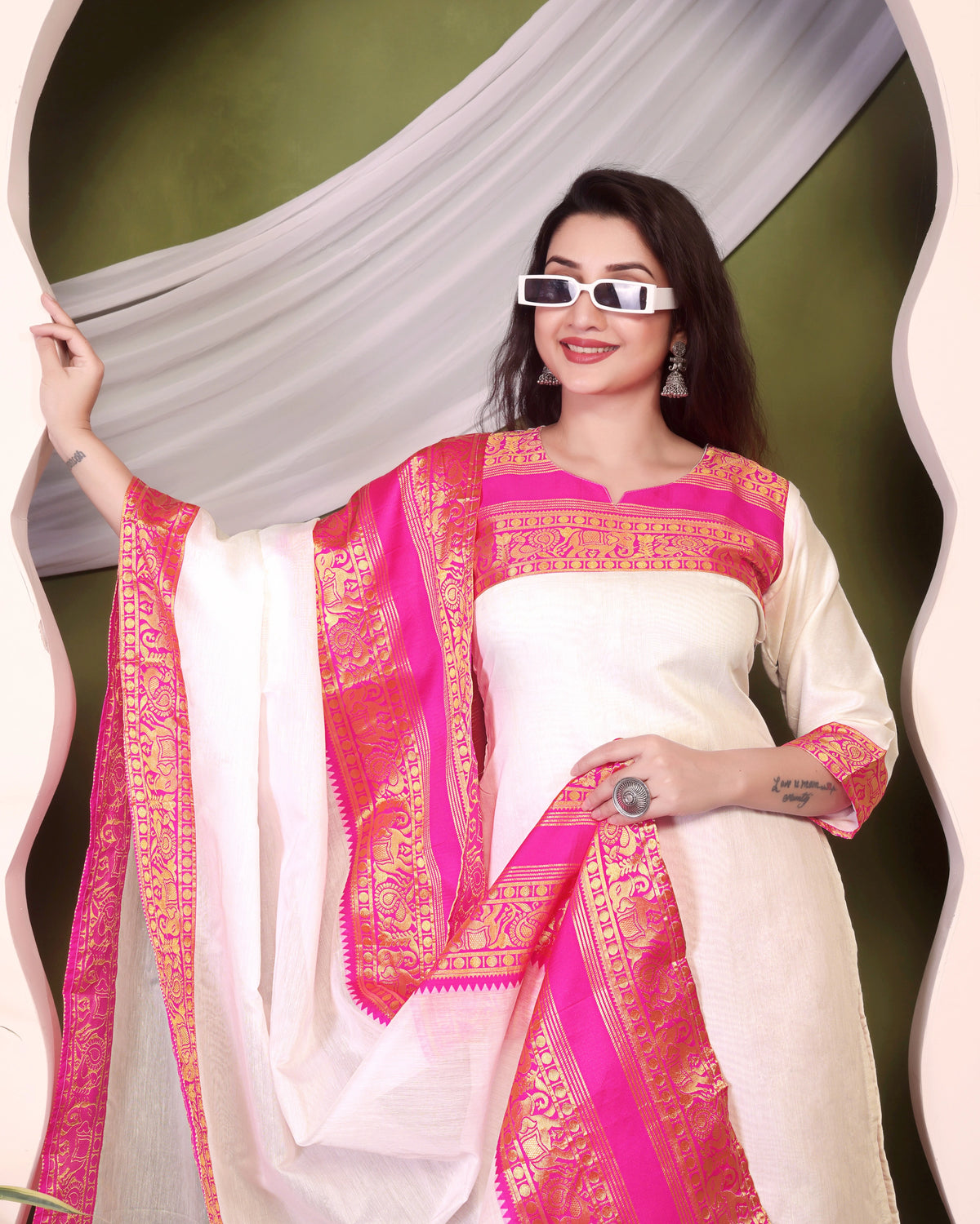 Woman wearing a white saree with pink and gold border against a green background