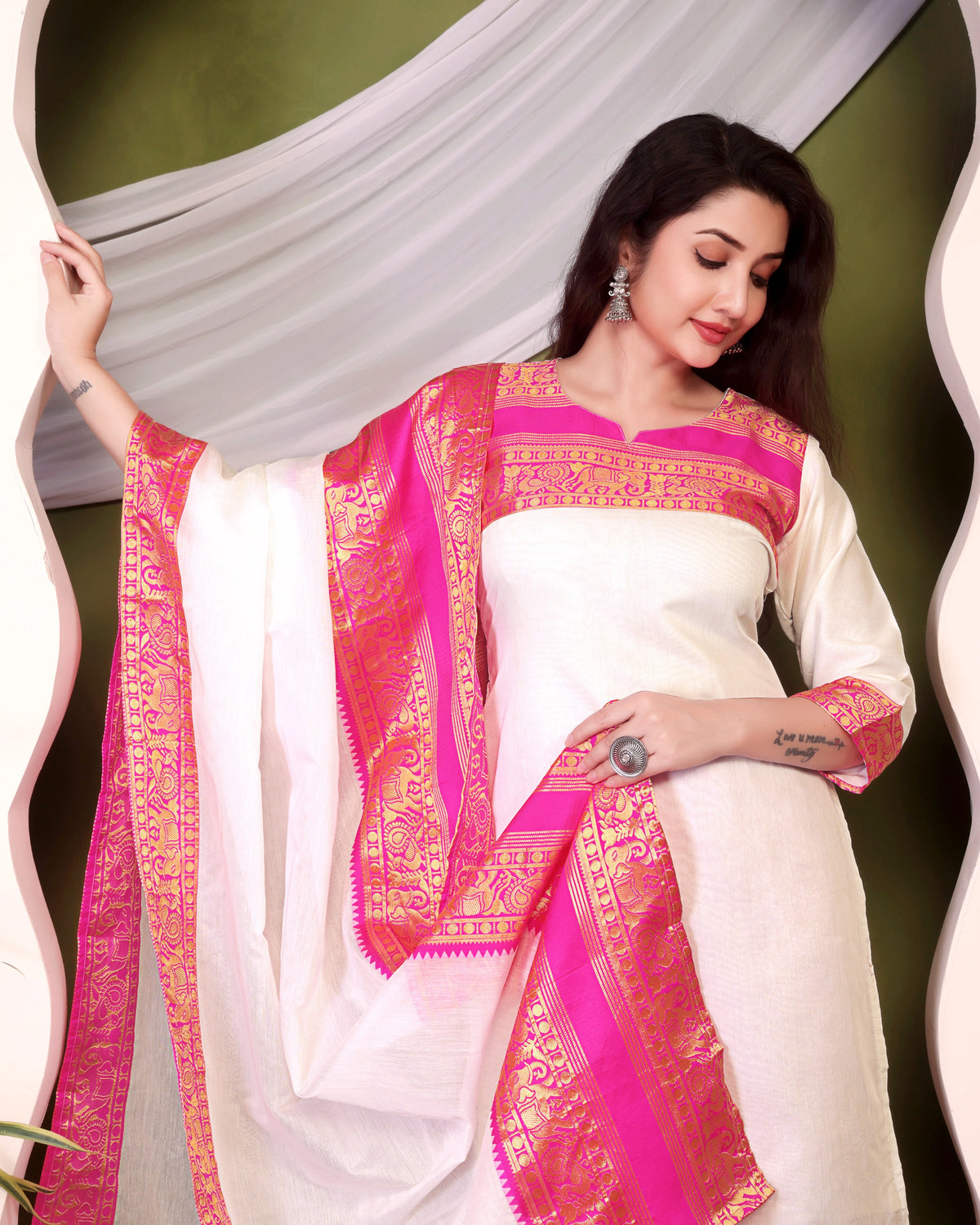Woman wearing a white saree with pink and gold border against a green background