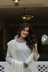 Woman in a white traditional outfit with intricate patterns, standing indoors.