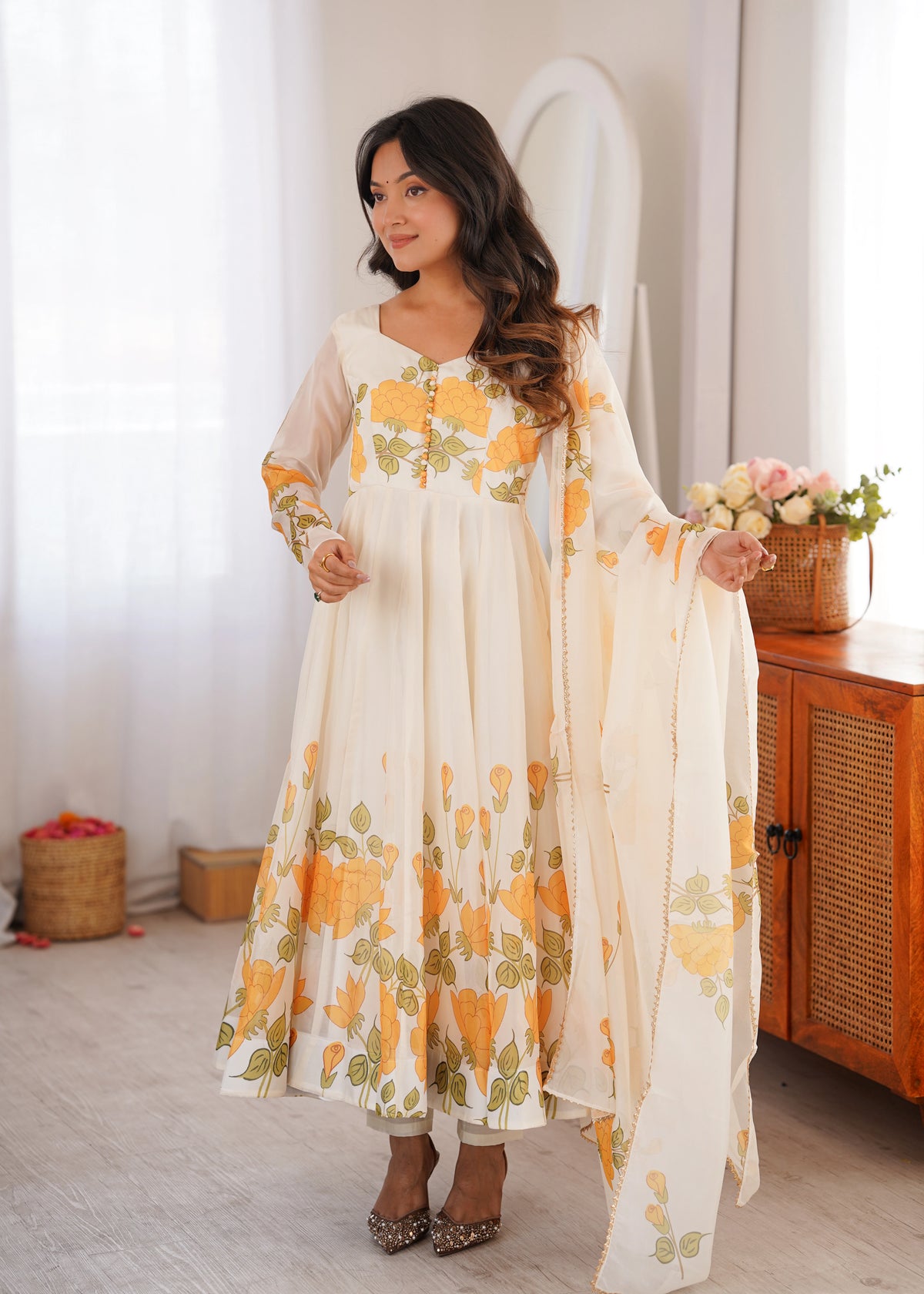 Woman wearing a floral dress standing in a room with wooden furniture and a white curtain.