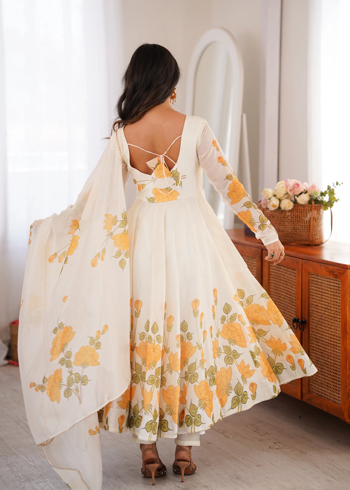Woman in a floral dress standing in a bright room with a wooden cabinet and flowers.