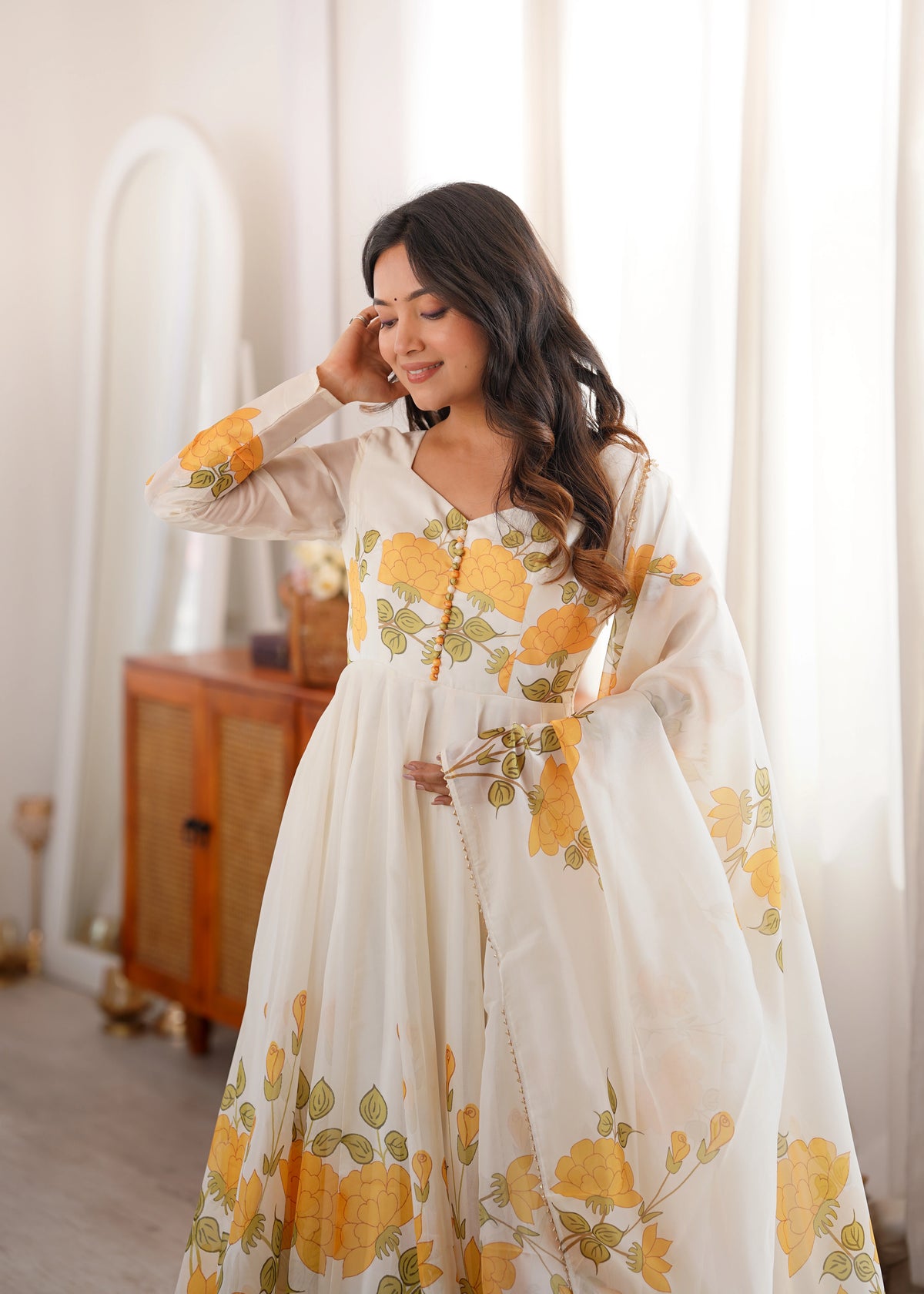 Woman wearing a floral dress with yellow flowers in a room with white curtains.