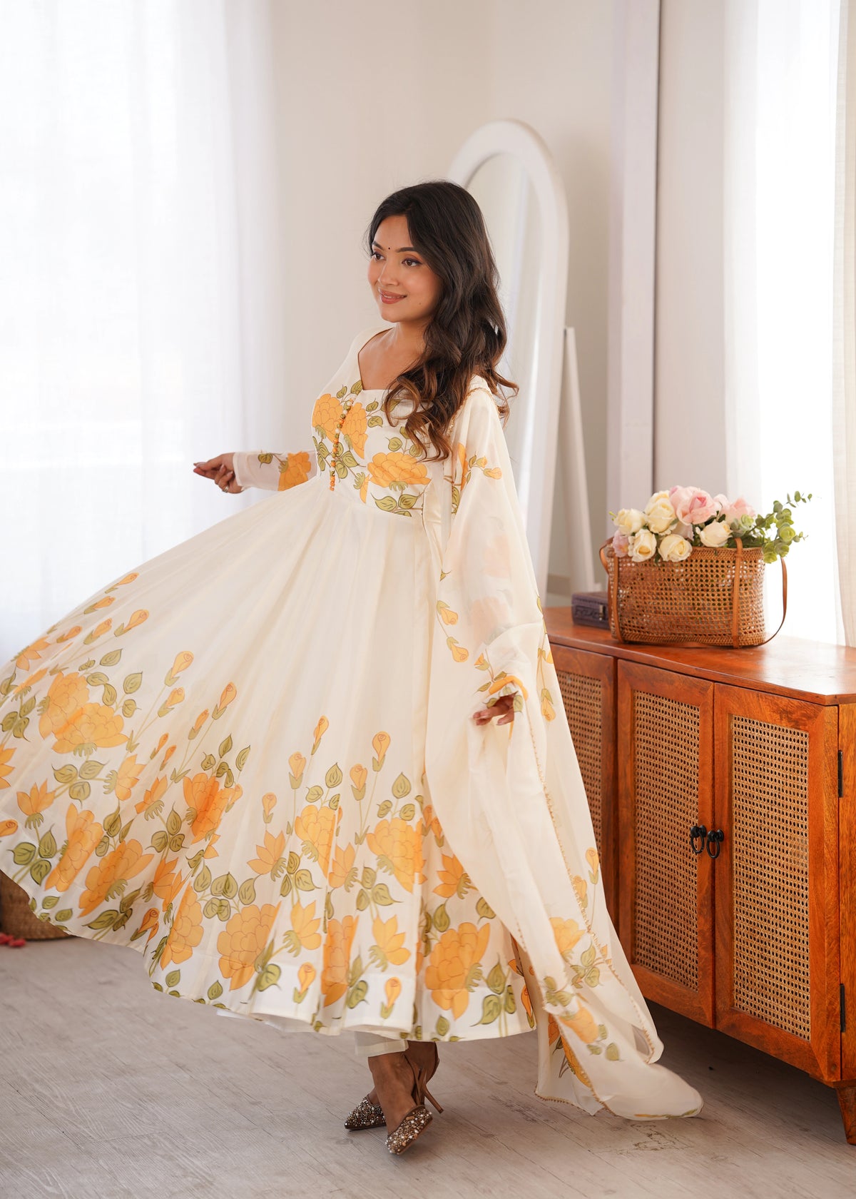 Woman in a floral dress standing in a room with a wooden cabinet and flowers.