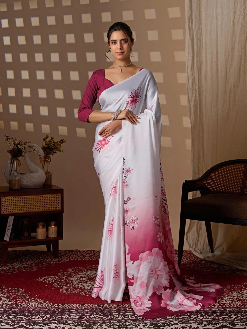 Woman wearing a white saree with pink floral patterns in a room with a checkered wall and a chair.