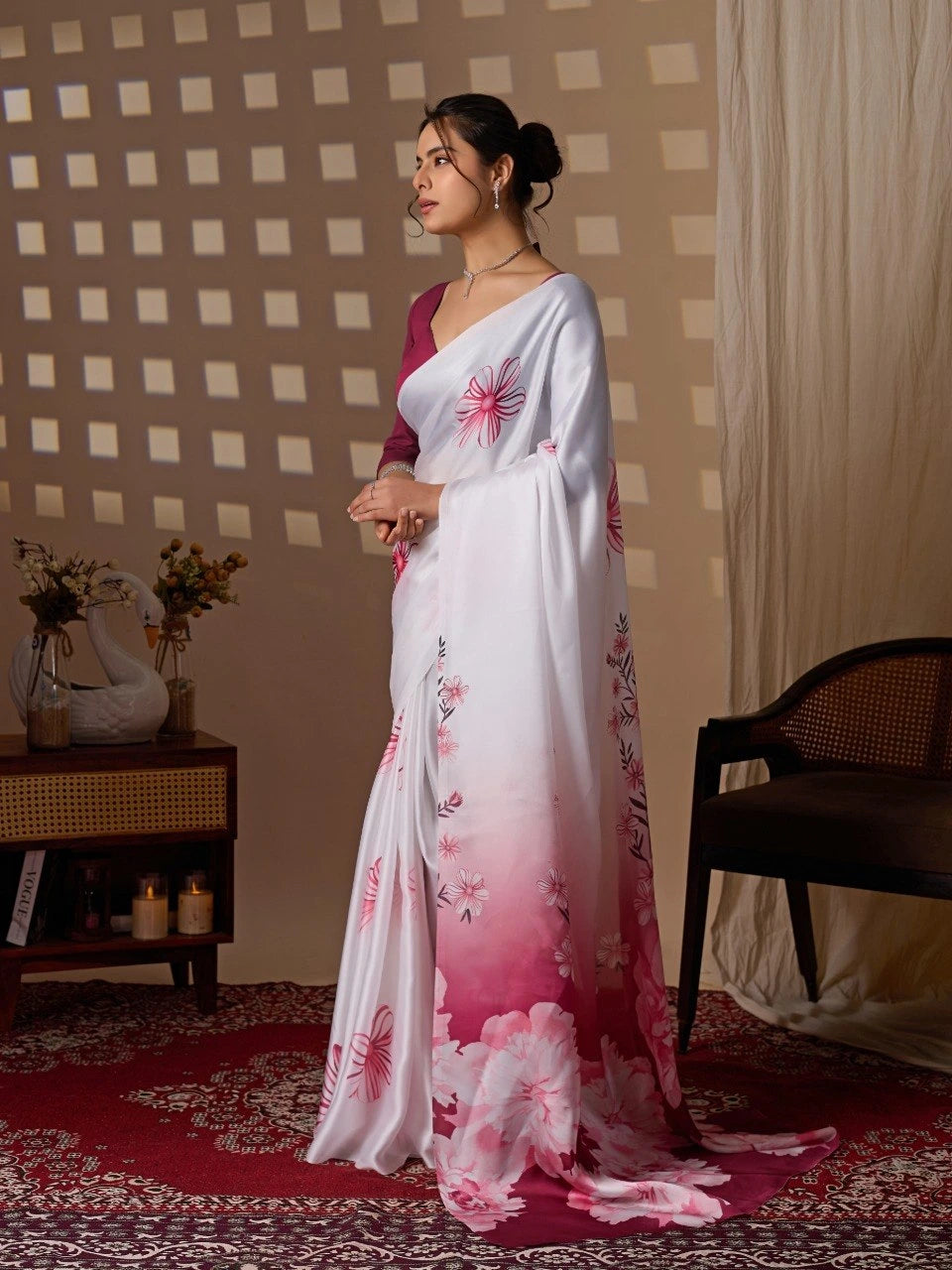 Woman in a white saree with pink floral patterns standing in a room with a checkered wall and wooden furniture.