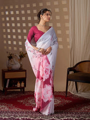 Woman in a white saree with pink floral patterns standing in a room with a checkered wall and a chair.
