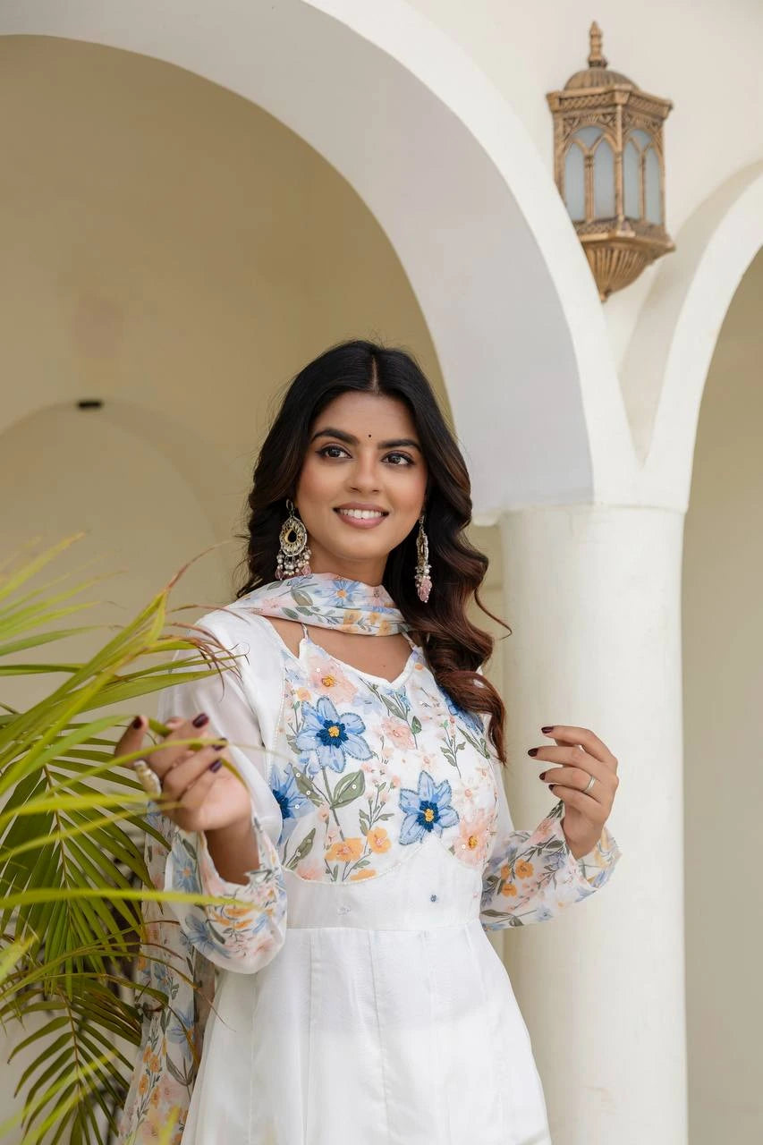 Woman in a white dress with floral embroidery holding a plant, standing in a decorative archway.