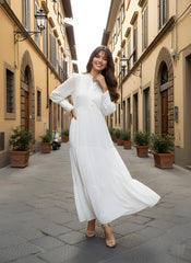 Woman in a white dress standing on a narrow street with buildings and potted plants.