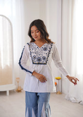 Woman wearing a white embroidered top with blue details in a bright room.