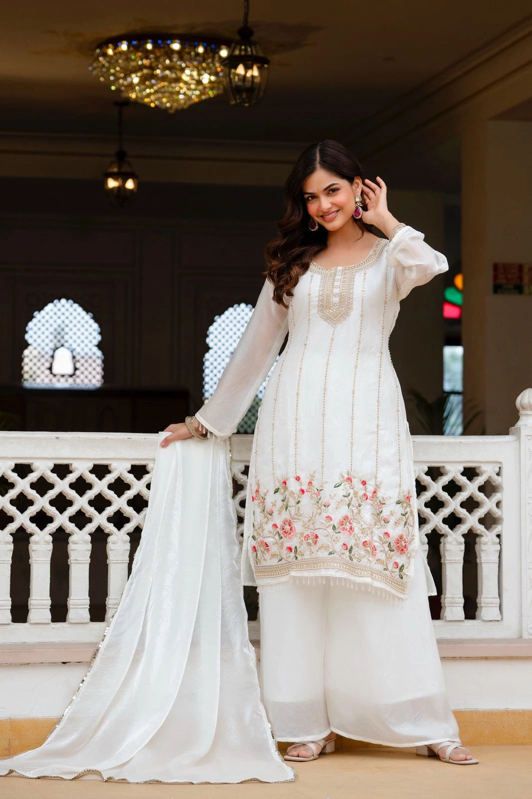 Woman in a white traditional outfit with floral embroidery standing indoors.