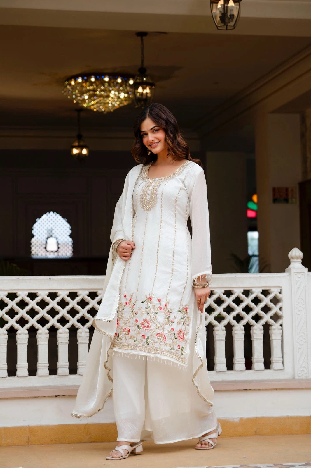 Woman in a white traditional outfit with floral embroidery standing in an elegant indoor setting.