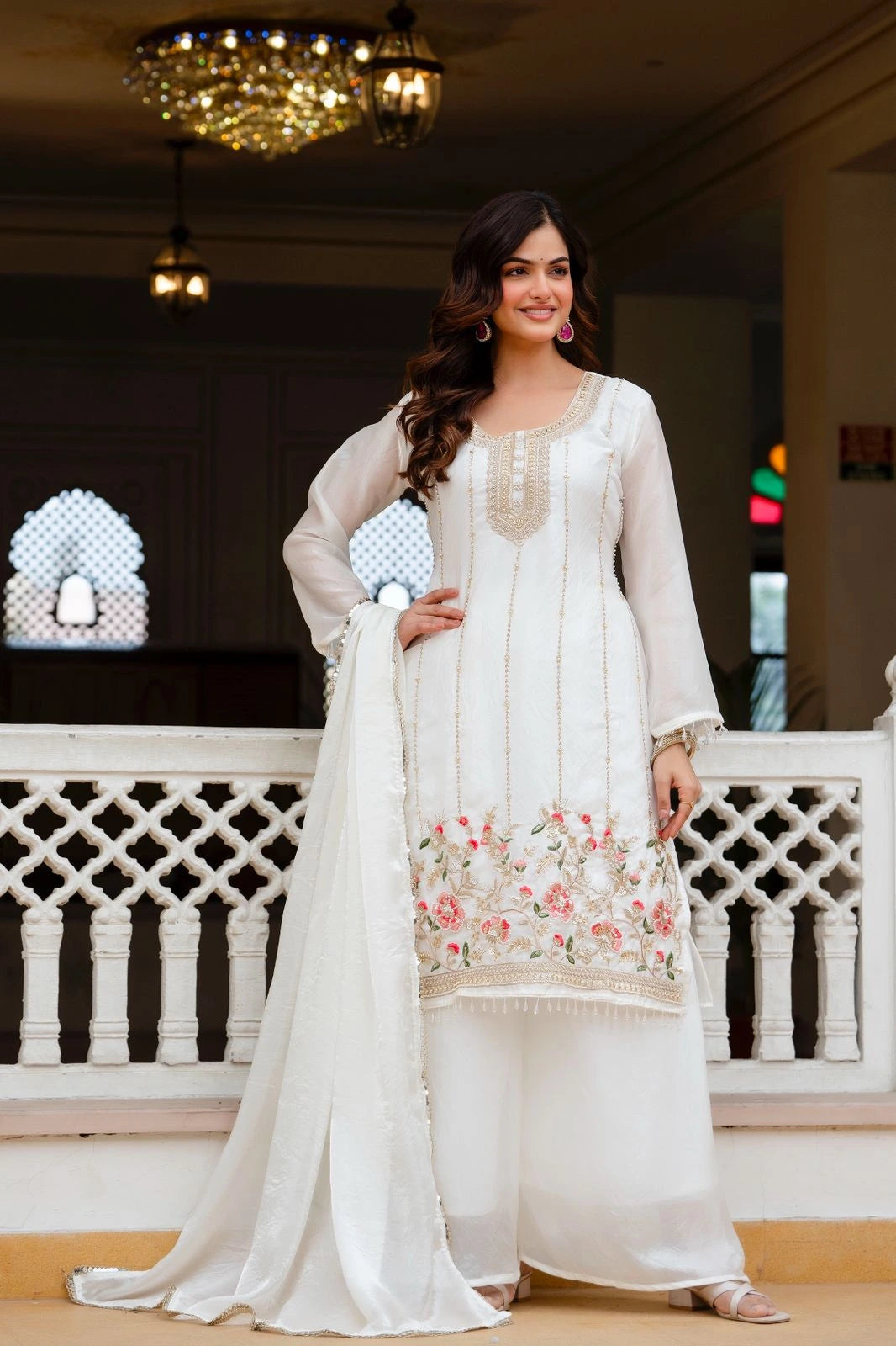 Woman in a white traditional outfit with floral design standing in an elegant indoor setting.