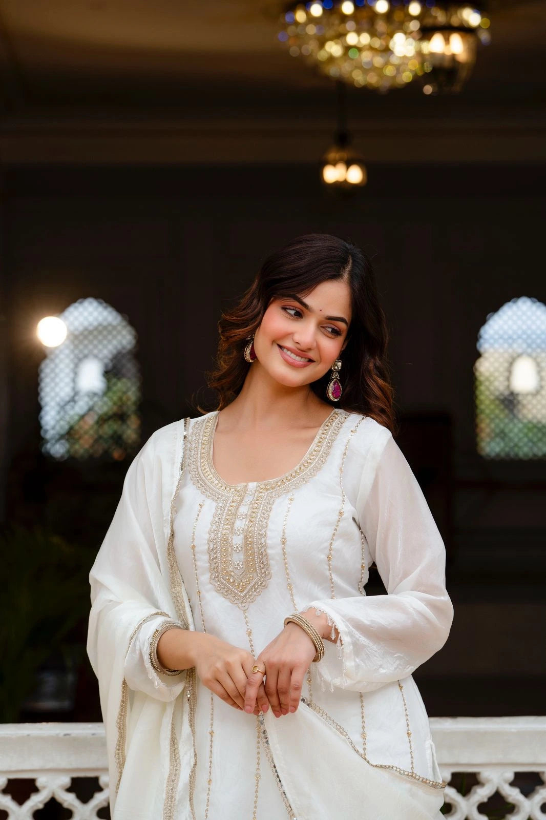 Woman in a white traditional outfit with intricate designs, standing in an elegant indoor setting.