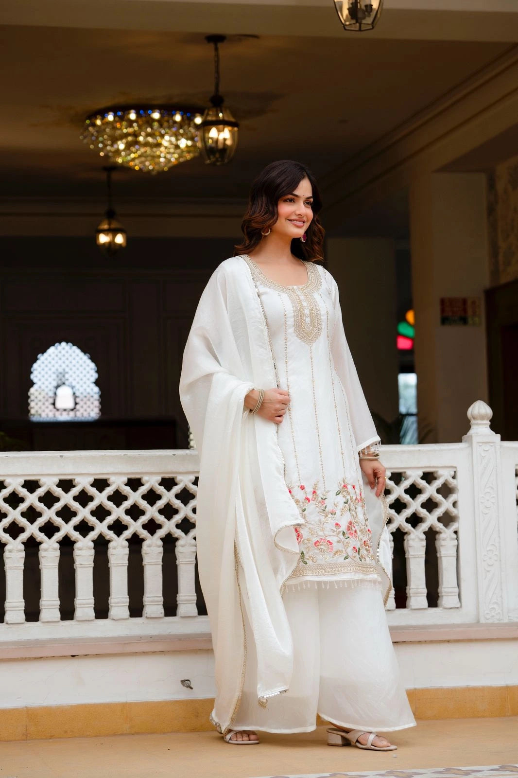 Woman in a white traditional outfit standing in an elegant indoor setting with chandeliers.