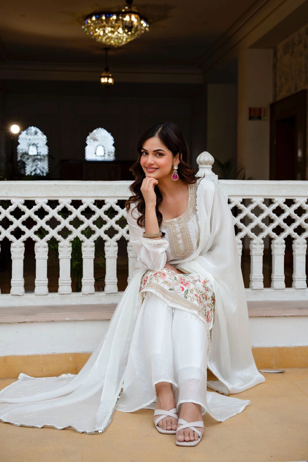 Woman in a white traditional outfit with floral patterns sitting on a balcony.