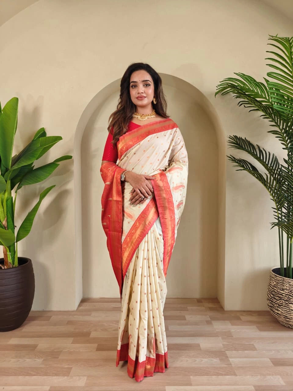 Woman in a red and white saree standing in a room with plants.