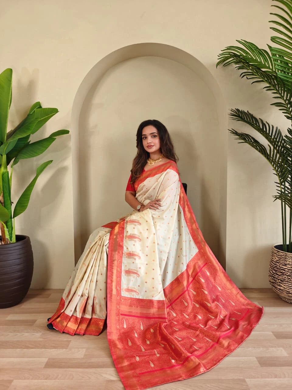 Woman in a white and red saree standing indoors with plants on either side.