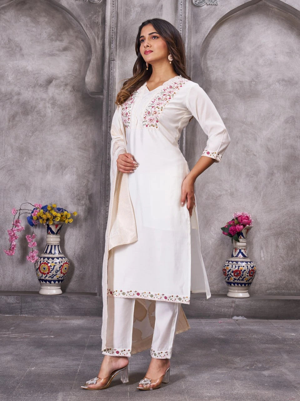 Woman in a white embroidered outfit standing in front of a gray wall with decorative vases.