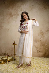 Woman in a white traditional outfit with a floral blouse, holding a white dupatta against a textured beige wall.