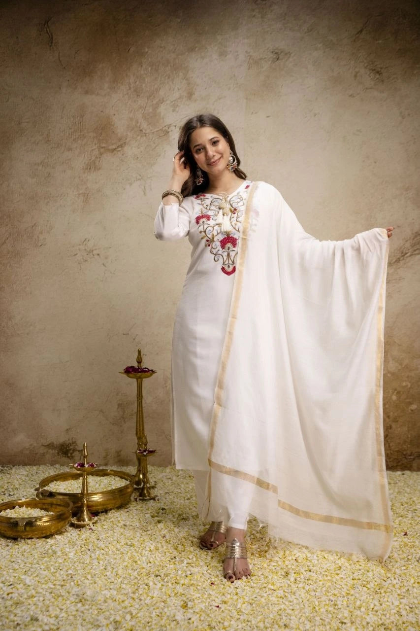 Woman in a white traditional outfit with floral embroidery standing against a textured wall.