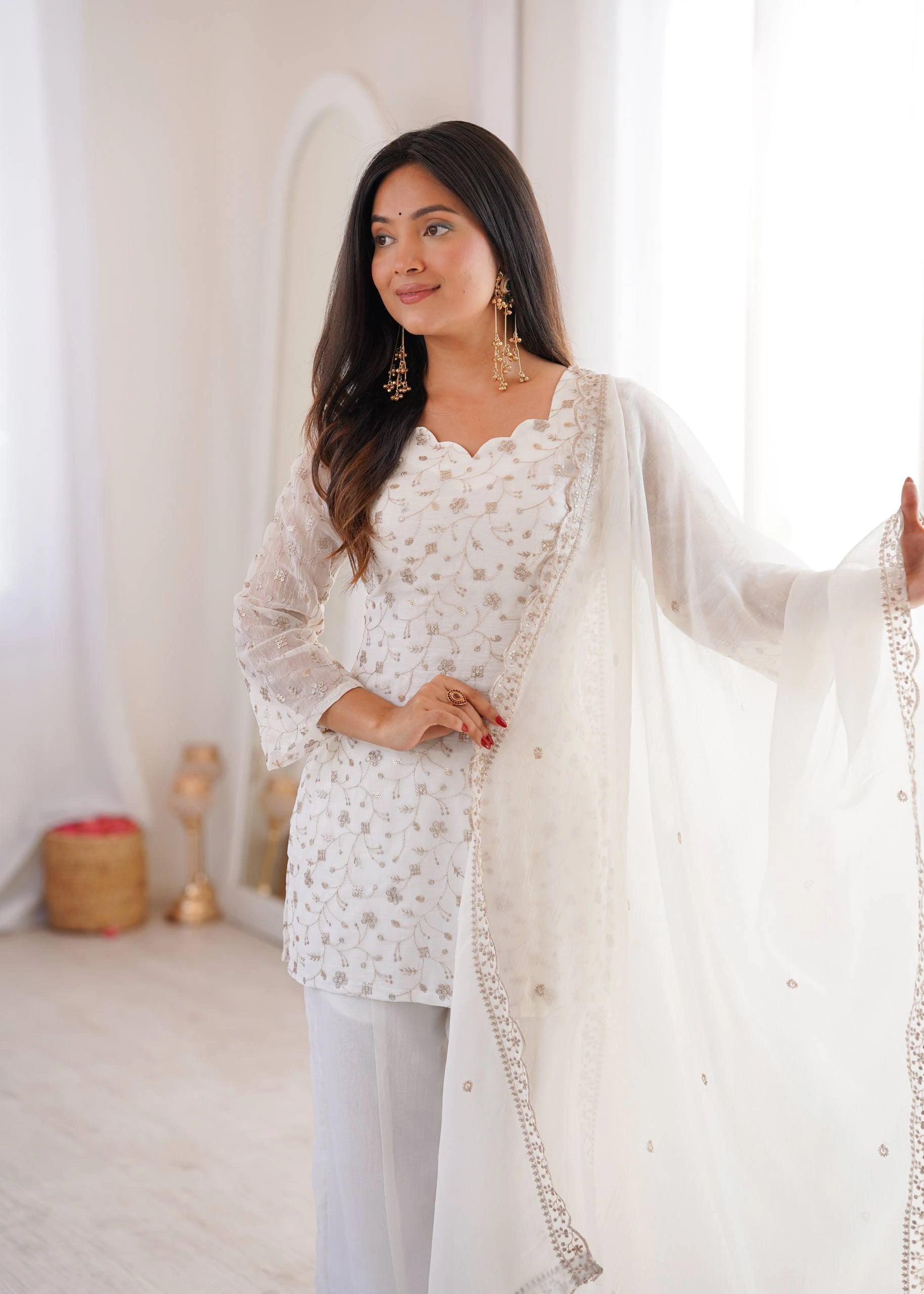Woman wearing a white embroidered outfit with a sheer dupatta in a softly lit room.