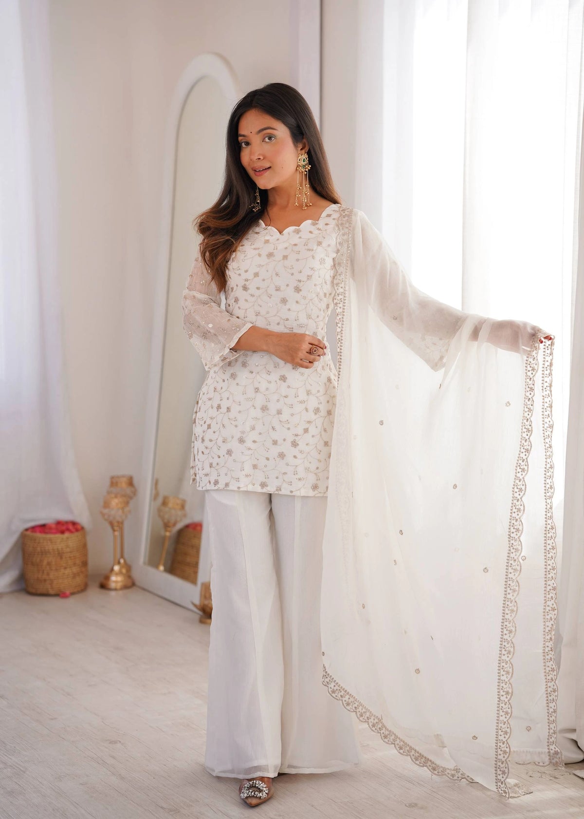 Woman in a white traditional outfit with a sheer dupatta, standing in a bright room.