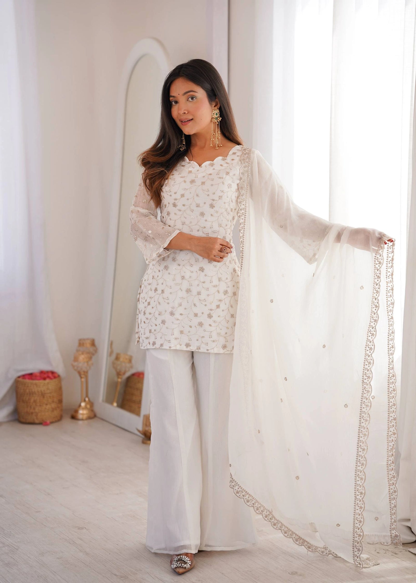 Woman in a white traditional outfit with a sheer dupatta, standing in a bright room.