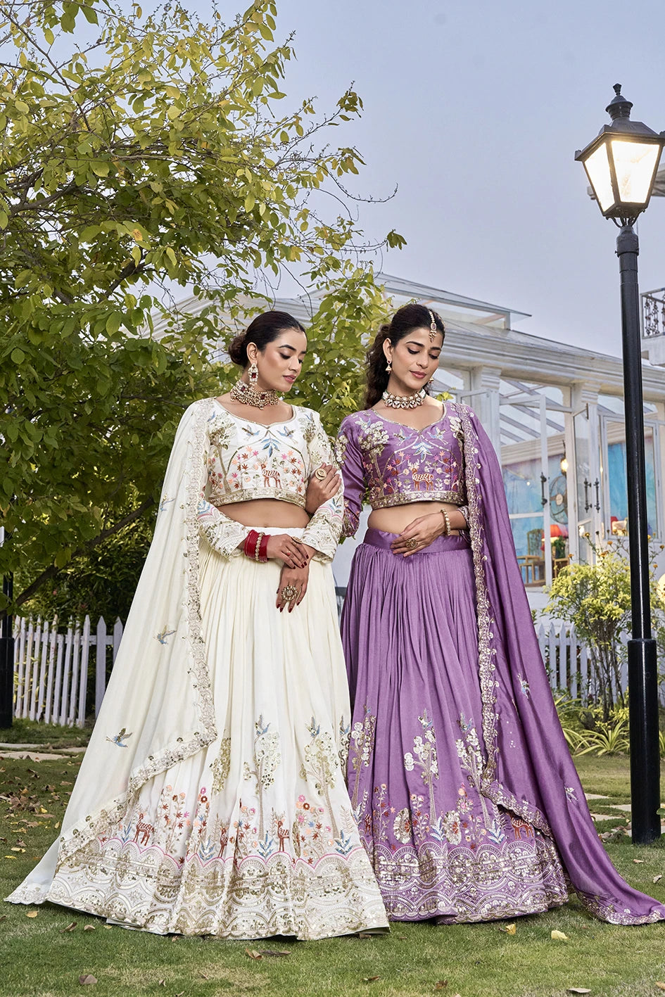 Two women in traditional embroidered dresses standing outdoors with greenery and a building in the background.