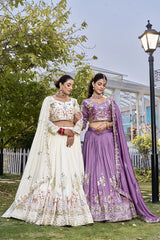 Two women in traditional embroidered dresses standing outdoors with greenery and a building in the background.