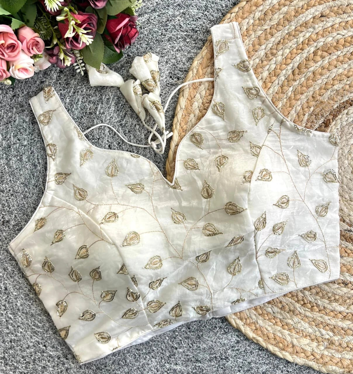 White blouse with gold patterns on a textured surface with flowers and a woven mat.