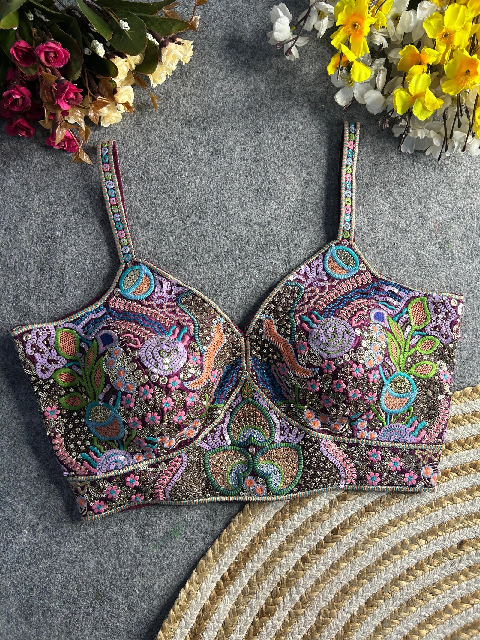 Traditional multicolor bustier-style blouse featuring intricate thread embroidery and sequin embellishments in floral and paisley motifs, with structured cups and adjustable straps, displayed flat on a textured grey surface surrounded by artificial pink, yellow, and white flowers.