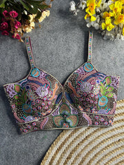 Traditional multicolor bustier-style blouse featuring intricate thread embroidery and sequin embellishments in floral and paisley motifs, with structured cups and adjustable straps, displayed flat on a textured grey surface surrounded by artificial pink, yellow, and white flowers.