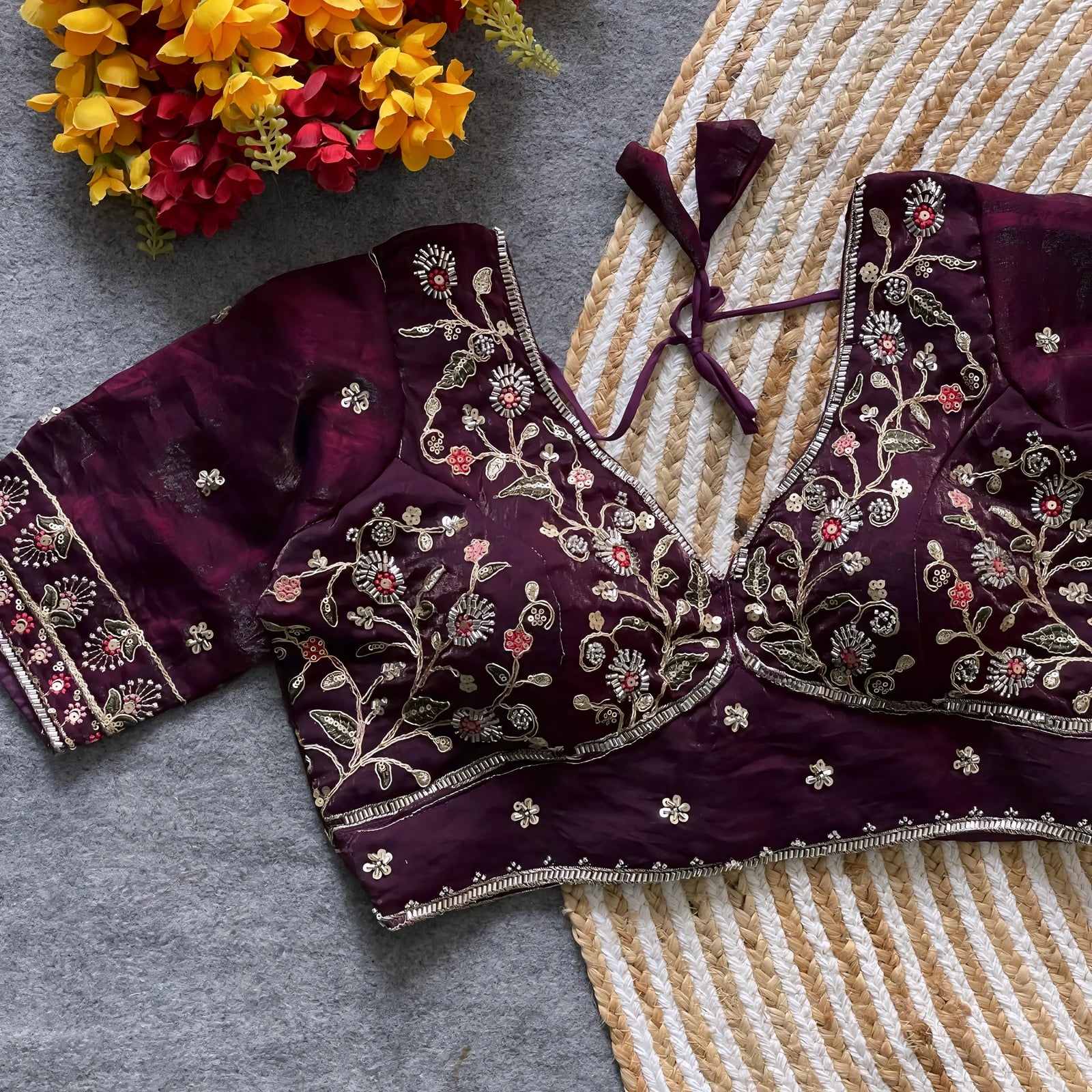 Wine padded blouse with intricate multicolor floral embroidery and sequin accents, featuring a tie-back detail, displayed on a grey surface with artificial yellow and red flowers and a woven mat in the background.