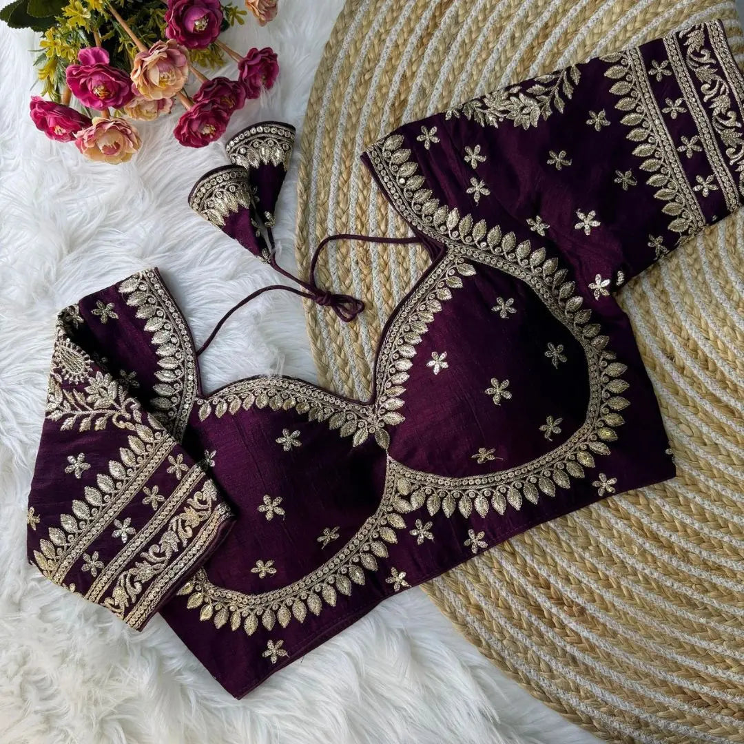 Maroon blouse with intricate gold zari embroidery featuring floral and leaf motifs, decorative borders on sleeves, and tie-up tassels, displayed on a textured mat with pink flowers in the background