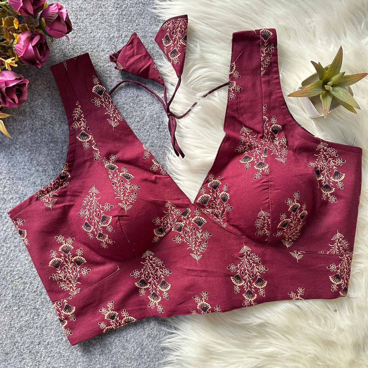 Maroon embroidered blouse with floral patterns on a textured surface with flowers and a plant.