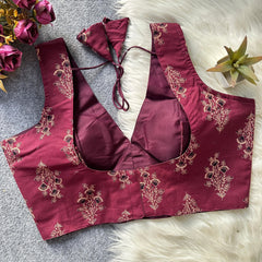 Maroon embroidered blouse with floral patterns on a textured surface with flowers and a plant.