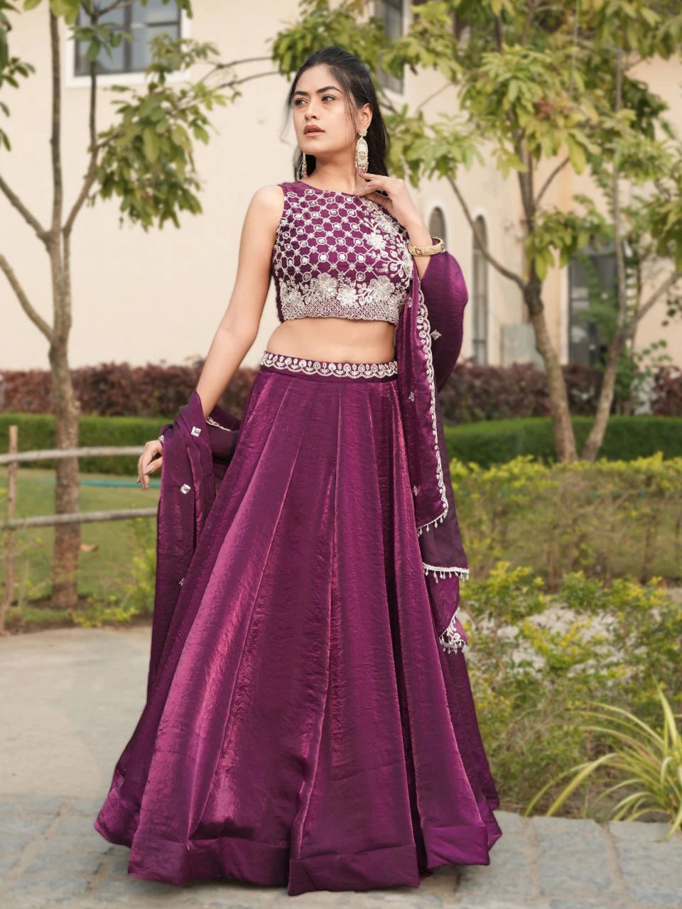 Woman in a purple traditional outfit standing outdoors with greenery in the background