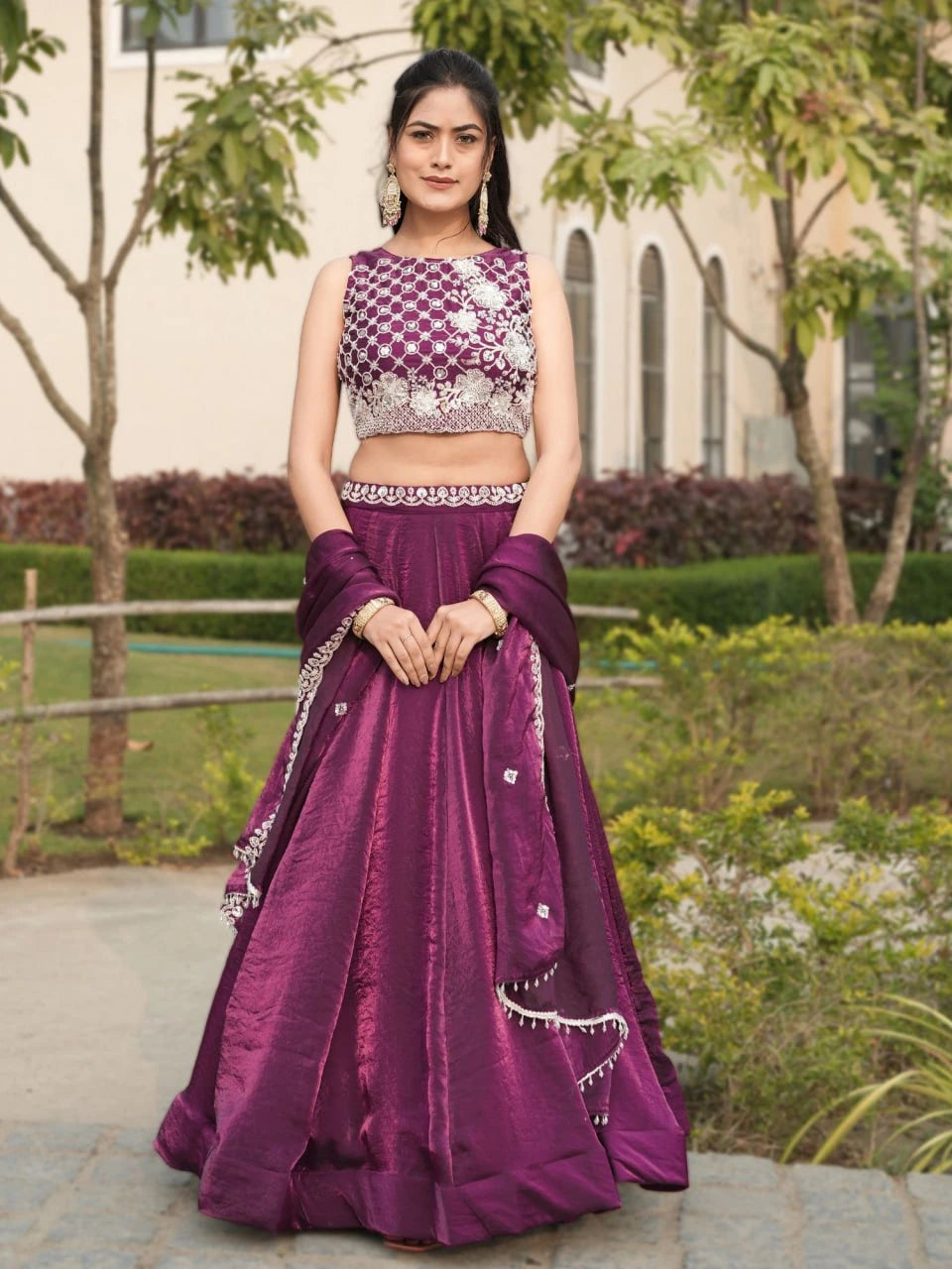 Woman in a purple traditional outfit standing outdoors with greenery in the background