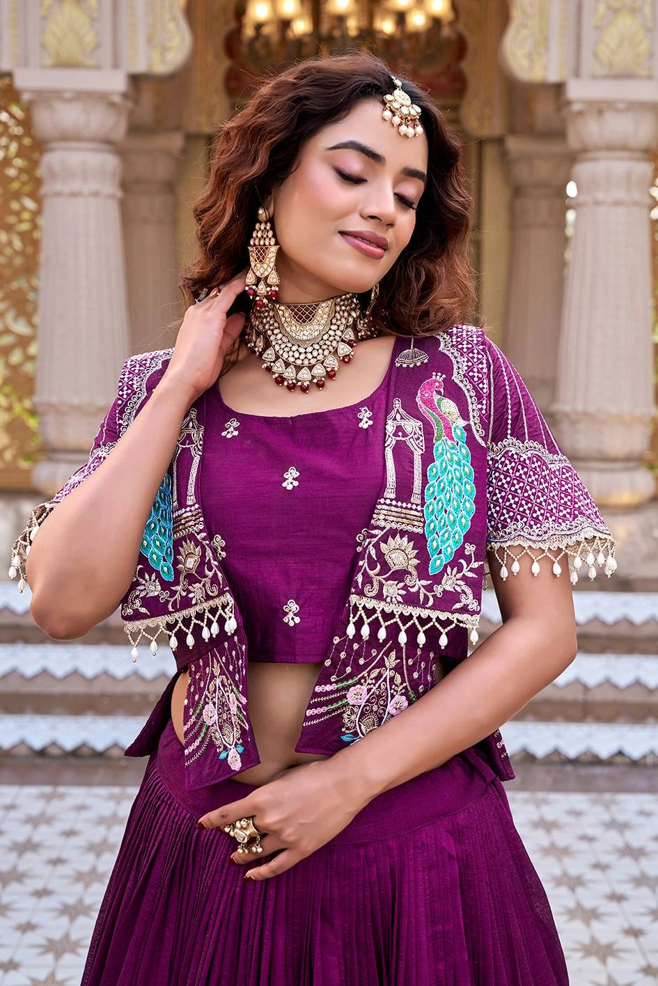 A waist-up portrait of the woman with her eyes softly closed, highlighting her traditional gold jewelry and the detailed patterns of the lehenga set. The background features ornate architectural pillars and a golden chandelier.