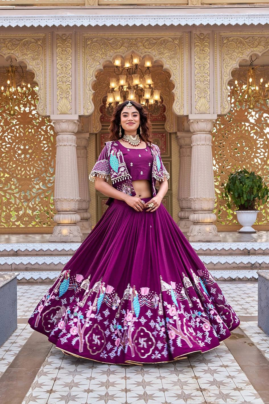 A full-length shot of a woman standing in a majestic courtyard wearing a deep magenta pleated silk lehenga. The skirt is heavily adorned with intricate peacock, floral, and traditional motifs in turquoise and pink. She is wearing a matching crop top and an open-front short jacket with pearl-fringe detailing.