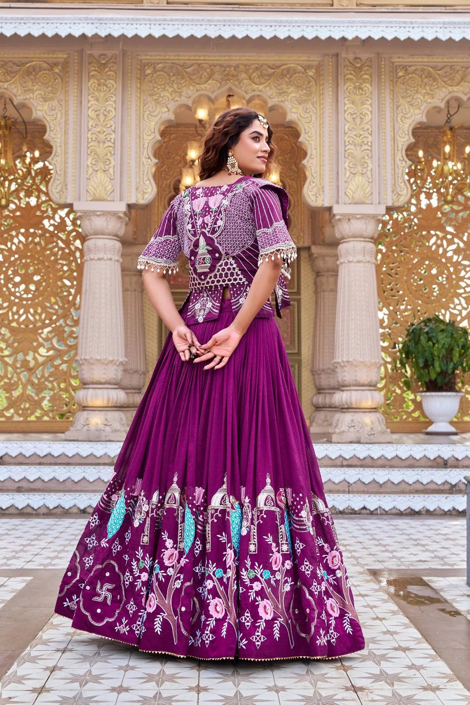 A full-length back view of the woman, showcasing the extensive embroidery on the back of the jacket, which features a central decorative vase motif. The photo also captures the full volume of the pleated magenta skirt and the repeating traditional patterns along the hemline.