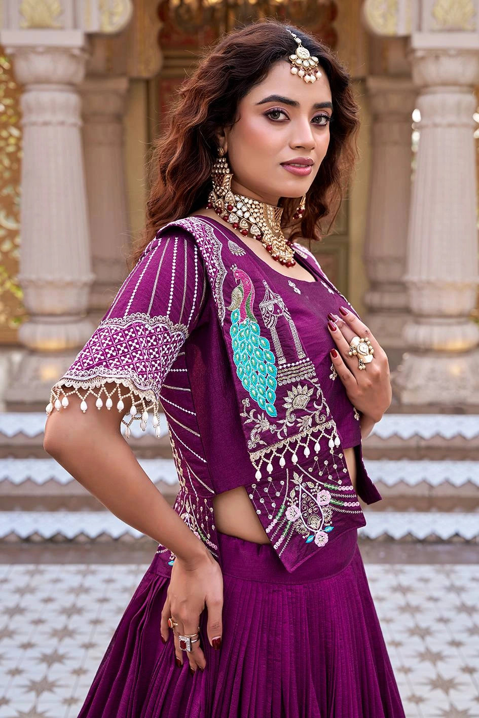 A medium close-up from the side, focusing on the craftsmanship of the magenta jacket. The embroidery features a vibrant turquoise peacock and elaborate gold threadwork, complemented by delicate pearl drops along the sleeve and hem. The woman is wearing a heavy kundan choker and matching earrings.