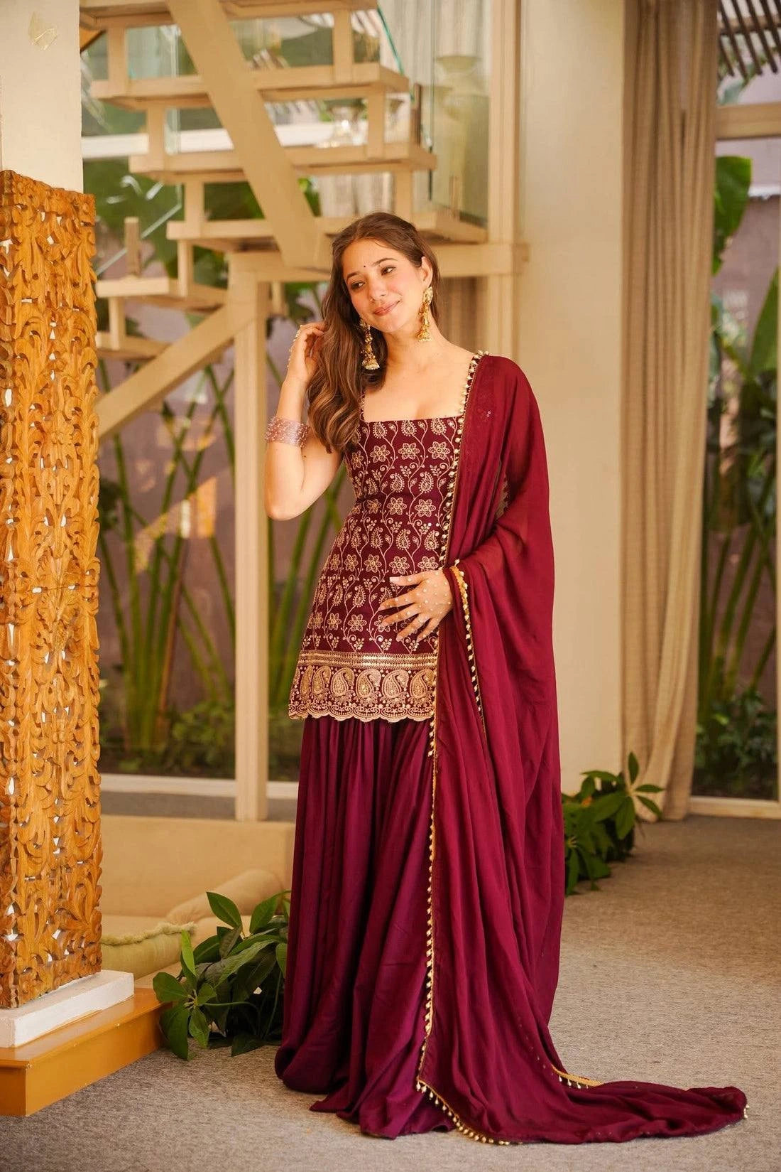 Woman in a burgundy traditional outfit standing in an indoor setting with plants and decor.