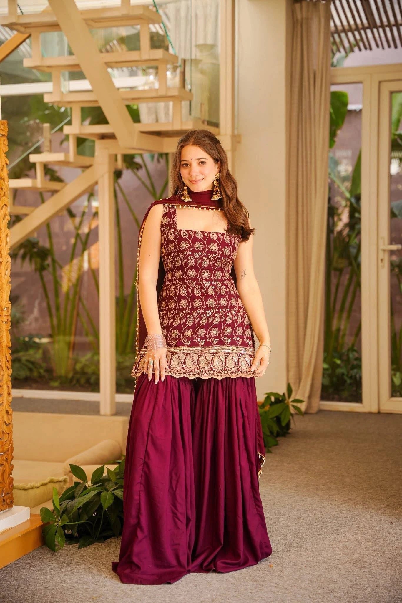 Woman in a maroon traditional outfit standing indoors with decorative elements.