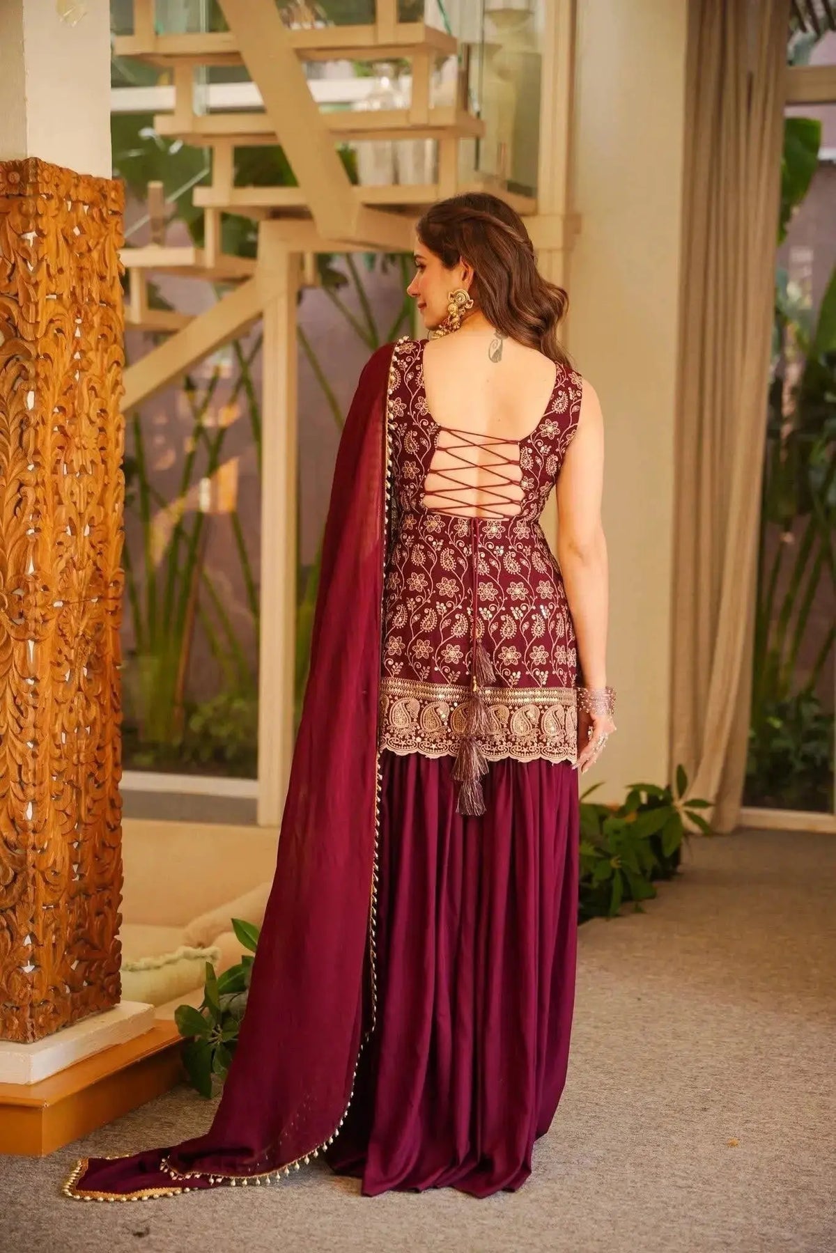 Woman in a maroon traditional outfit with intricate designs, standing indoors.