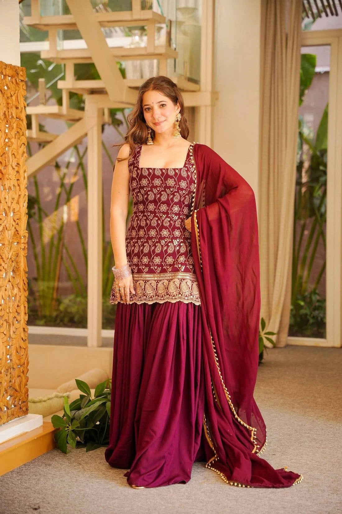 Woman in a traditional maroon and gold outfit standing indoors with decorative elements.