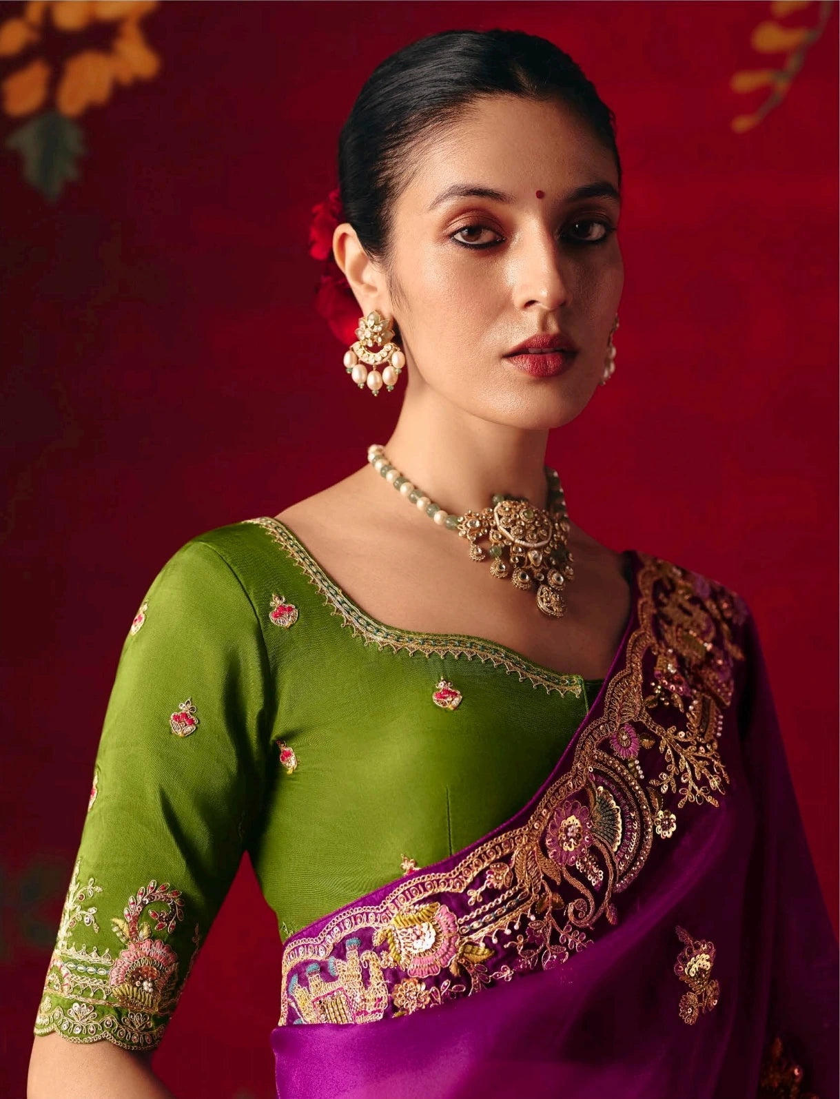 Woman wearing a green blouse with floral patterns and a purple saree with gold embroidery against a red background.