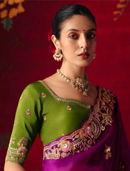 Woman wearing a green blouse with floral patterns and a purple saree with gold embroidery against a red background.