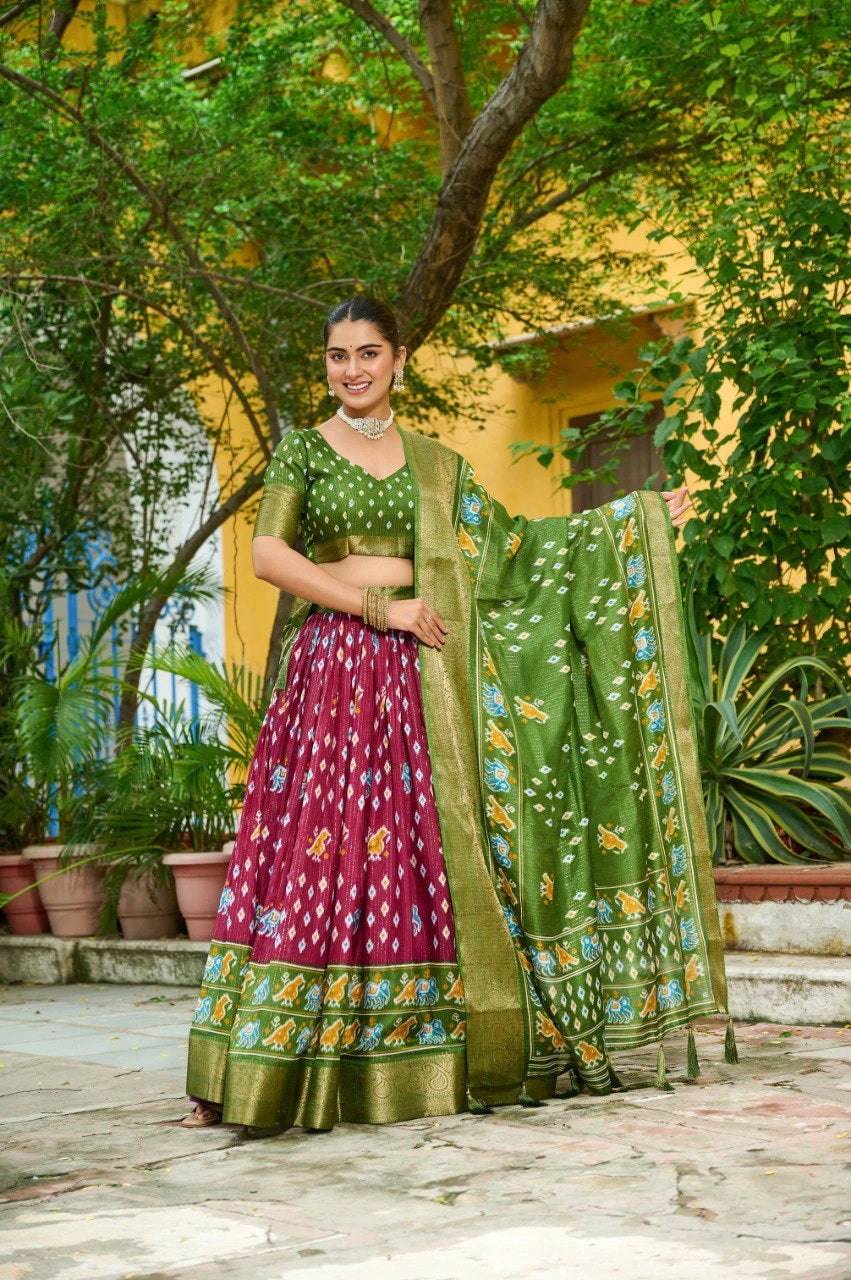Woman in a green and pink traditional outfit standing outdoors with trees and plants in the background